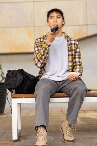 Young asian man talking into phone while eating sandwich on urban bench during lunch break