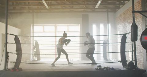 Sparring coach training female trainee in backlit loft boxing ring, warehouse gym