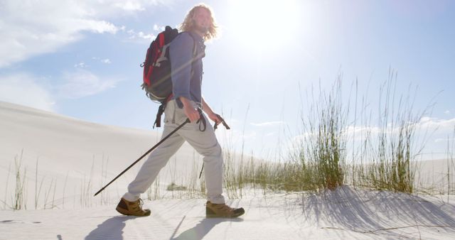 Blonde man hiking through a sandy desert under bright sunlight. Wearing a backpack, holding hiking poles, partially shaded by long plants. Suitable for themes related to adventure, exploration, outdoor activities, nature travel, trekking or desert environments.