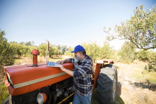 Farmer using laptop on tractor in olive farm on a sunny day. Ideal for illustrating modern farming practices, agricultural technology, and rural life. Can be used in articles about farming innovations, digital tools in agriculture, or rural business management.