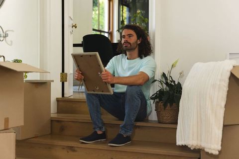 Man sitting on wooden steps unpacking boxes holding picture frame in sunlit foyer