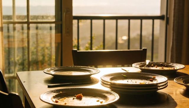 Golden-hour sunlight streaming over stacked plates with crumbs on wooden dining table