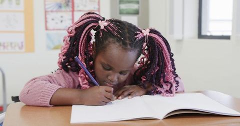 Young girl with colorful braids focused on writing in classroom