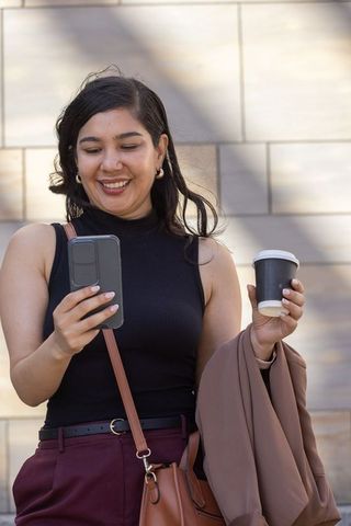 Smiling woman checking smartphone holding coffee and blazer on sunlit stone facade