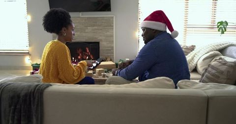 Cozy Couple Wrapping Holiday Gifts by Fireplace While Using Tablet and Wearing Santa Hat