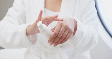 Indian woman applying moisturizer from white pump bottle while wearing plush white bathrobe
