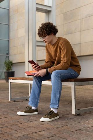 Teen student checking phone while studying on bench in modern plaza with notebook and pen