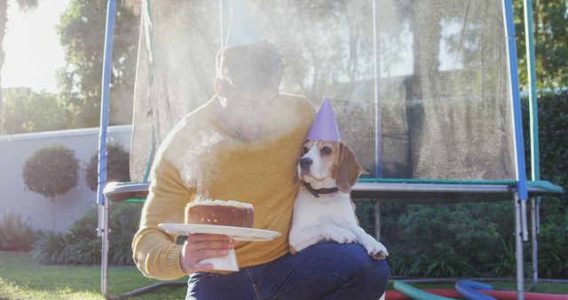 A Caucasian man celebrates a birthday outdoors with his beagle, both wearing party hats and a cake in hand, with copy space. Their shared moment highlights a joyful pet-owner relationship and the inclusion of pets in family celebrations.
