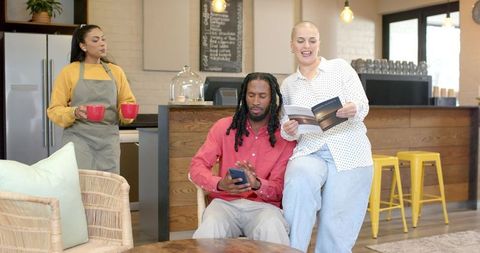 Barista serving red mugs while patrons relaxing, reading and using smartphone in cafe lounge