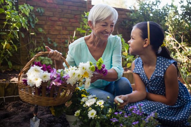 Elderly woman smiling while holding a basket of flowers conversing with young girl in backyard. Ideal for themes of family bonding, nature activities, and gardening. Great for use in advertisements promoting family time, gardening, and lifestyle brands.