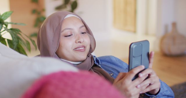 Image of smiling biracial woman in hijab using smartphone relaxing on sofa in living room at home. Happiness, communication, relaxation, inclusivity and domestic life.
