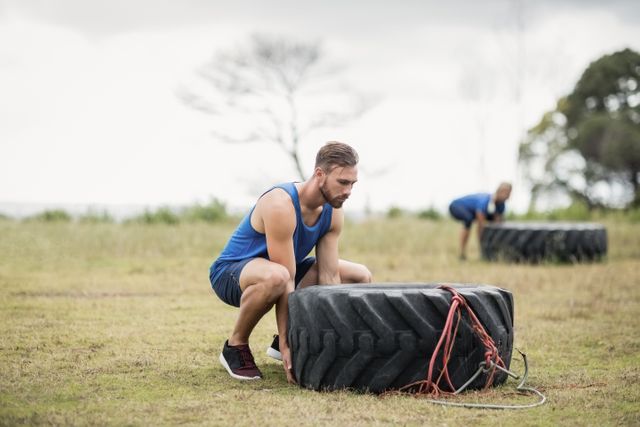 Man flipping a large tire in an outdoor boot camp setting. Ideal for use in fitness and exercise promotions, strength training programs, outdoor workout advertisements, and athletic training materials.