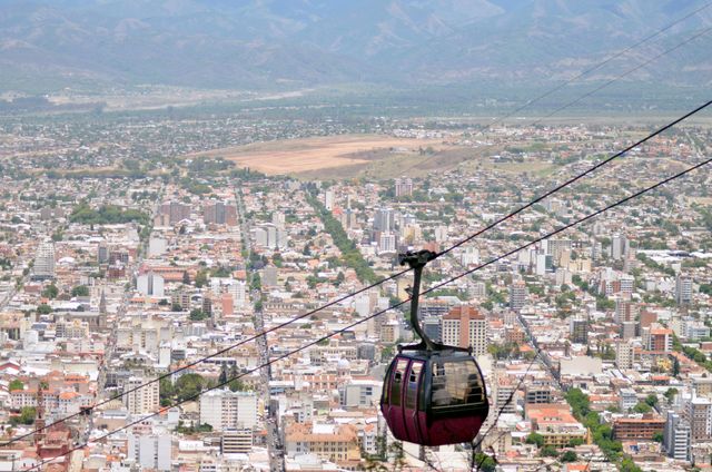 A scenic aerial view showcasing an urban setting with a cable car moving across above the city. The landscape includes numerous buildings, stretching into the distance and framed by mountainous terrain. Ideal for use in travel blogs, city guides, and tourism promotions depicting urban adventure and innovative transportation.