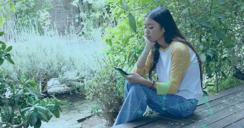 Young Asian woman sitting on deck using smartphone, contemplative in green courtyard with AR overlay