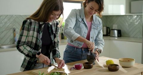 Two women preparing meal chopping red onion and grinding herbs on sunlit kitchen island