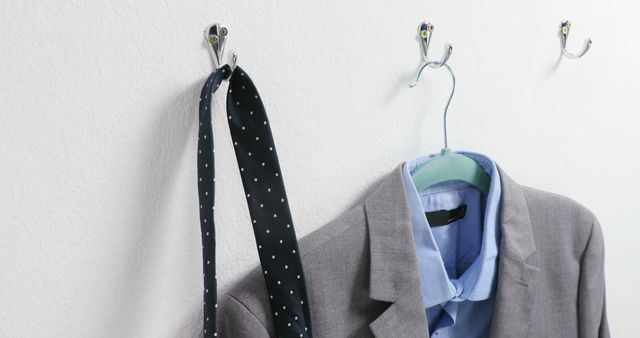 A gray business suit jacket and a polka-dotted tie hang on wall hooks against a white background, with copy space. This setup suggests a professional environment, preparing for a workday or a formal event.