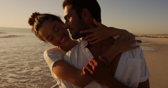 Biracial couple embraces lovingly on a beach at sunset. Their affectionate pose captures a romantic moment by the sea.