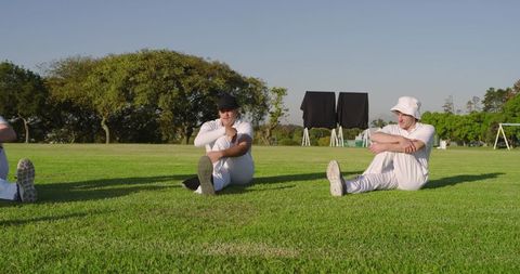 Cricket players stretching on outfield in white uniforms during warmup under sunny sky