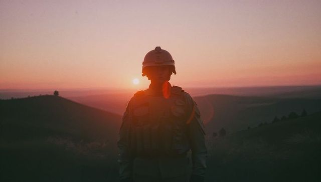 Solitary soldier in combat gear at sunset on grassy hilltop