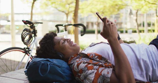 Young man enjoying leisure time lying down in an urban park on a sunny day, smiling while using smartphone. Wearing headphones and casual attire. Bicycle in the background. Ideal for promoting lifestyle, social media, mobile technology, relaxation, and outdoor activities.