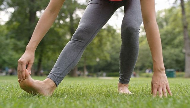 Woman stretching forward toe touch holding foot on grassy park lawn barefoot