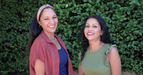 Smiling african american and indian women side-by-side by green hedge celebrating friendship