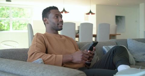 Relaxed african american man lounging on sofa holding smartphone in modern open-plan living room