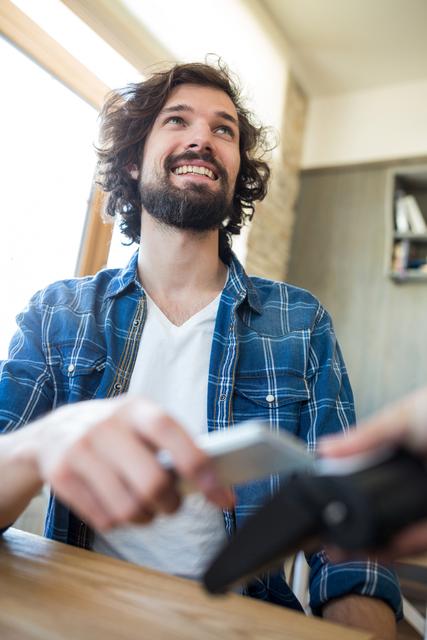 Young man in casual attire smiling while making a contactless payment with his mobile phone in a coffee shop. Ideal for illustrating modern payment methods, digital transactions, and lifestyle themes in marketing materials, blogs, or advertisements.