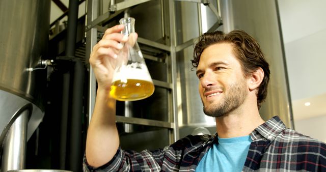 Man in casual attire inspecting beer in beaker inside brewery. Ideal for use in articles about the brewing process, craft beer manufacture, quality control in beverage industry or showcasing brewing professions.