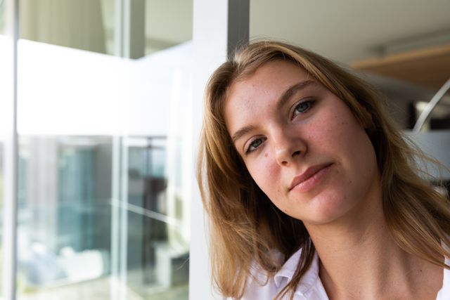 Portrait of a young Caucasian woman wearing a white shirt, standing on a balcony, looking at the camera, with her apartment in the background