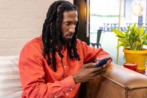 African american man relaxing on brown leather sofa using smartphone in bright cafe
