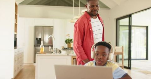 African American father watching son using laptop at modern open-plan kitchen island