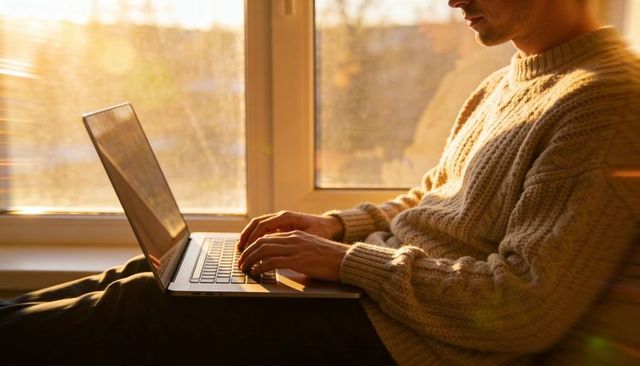 Man typing on laptop in cable knit sweater by sunlit window for remote work and cozy lifestyle