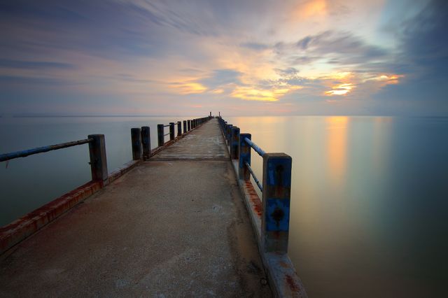 Photograph of a long pier extending into a calm ocean during sunset. The colorful sky reflects on the tranquil water, creating a peaceful and serene atmosphere. Useful for travel blogs, websites about relaxation and tranquility, wallpapers, and meditation guides.