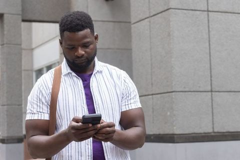 Young african man texting on smartphone on urban sidewalk by stone-clad building