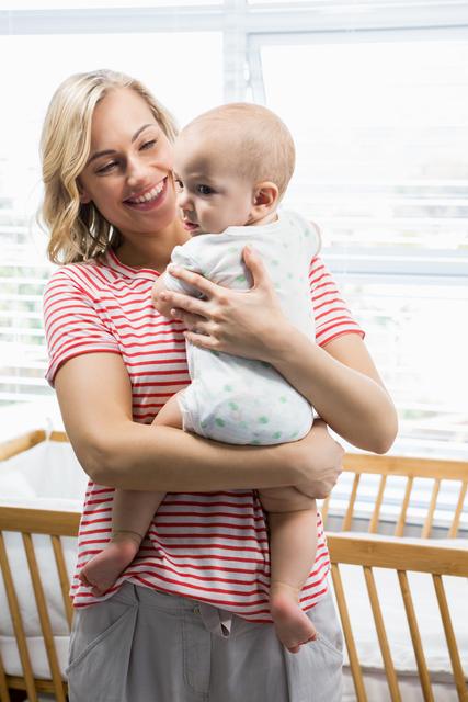 A mother is holding her baby boy while smiling at home. Ideal for use in parenting blogs, family magazines, and advertisements promoting childcare products, family services, or home living environments.