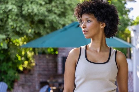 African american woman standing on sunlit backyard patio with teal umbrella, calm gaze