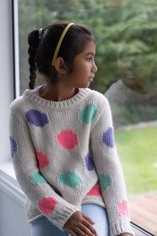 School-age asian girl sitting on window ledge gazing outside in cozy polka-dot sweater