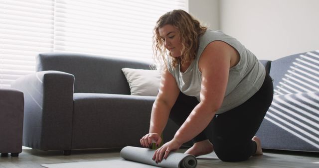 Curly-haired woman wearing casual clothing rolling up a yoga mat in a modern, minimalistic living room after a home workout session. Great for promoting home fitness routines, wellness blogs, or yoga equipment. Depicts a healthy lifestyle and determination.