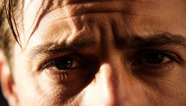 Holding gaze closeup male face showing furrowed brow stubble under warm dramatic light