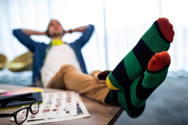 Casual man wearing colorful socks is relaxing with his feet on the desk in a modern office. This image can be used to depict workplace relaxation, work-life balance, and a laid-back office culture. It is ideal for articles or advertisements related to modern work environments, employee well-being, and office lifestyle.