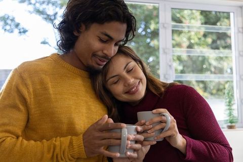 Couple sharing cozy morning coffee by window wearing knit sweaters and smiling