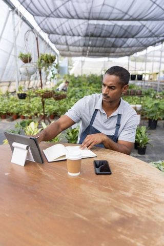 Indian nursery owner sitting at greenhouse table using tablet and notebook for inventory