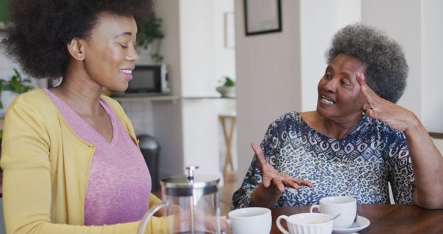 Young woman and elderly woman having a friendly talk while drinking coffee, creating warm atmosphere in kitchen. Perfect for depicting family bonds, friendship, intergenerational relationships, and moments of relaxation and joy.