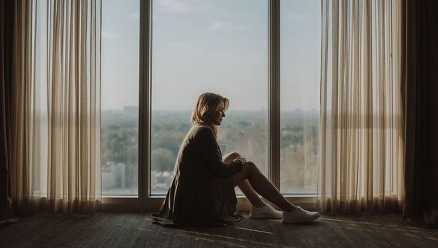 Sitting woman looking out floor-to-ceiling window over highrise treeline, sunlit contemplative silho