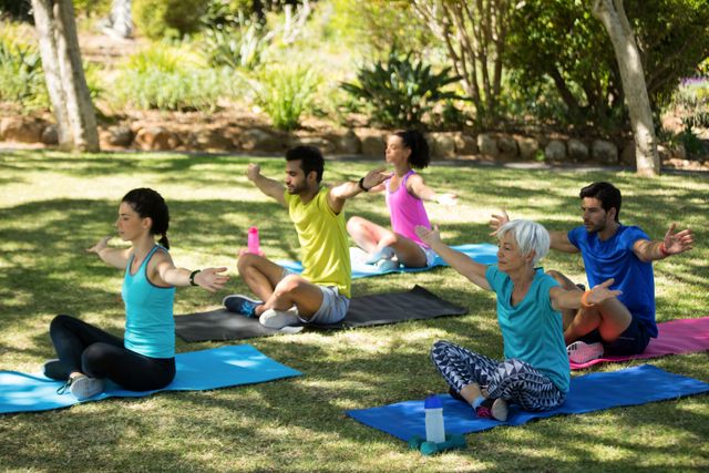 Group of people sitting on yoga mats with arms extended, practicing yoga in a park on a sunny day. This can be used for promoting outdoor fitness classes, health and wellness blogs, advertisements for yoga gear, or community wellness programs.
