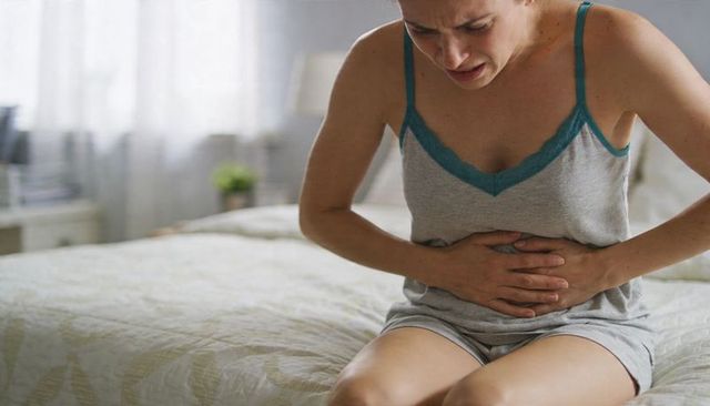Woman clutching lower abdomen on bed in grey cami sleepwear experiencing abdominal pain