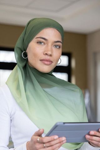 Confident woman wearing green hijab holding tablet in modern office portrait