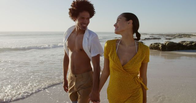 Biracial couple enjoys a romantic walk on the beach. Their carefree stroll by the sea epitomizes a perfect summer getaway.