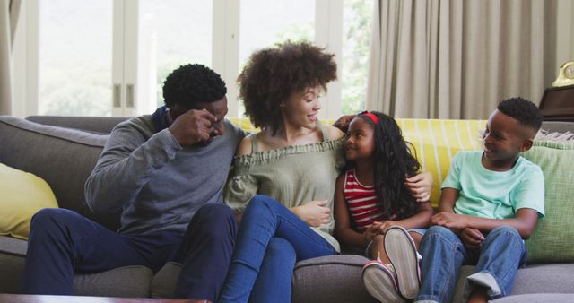 Parents and their two children are sitting together on a couch, enjoying their time with each other. The natural lighting and cozy environment suggest a warm and loving home setting, making this photo ideal for illustrating family bonds, home living, child-parent relationships, or promoting family-friendly products and services.